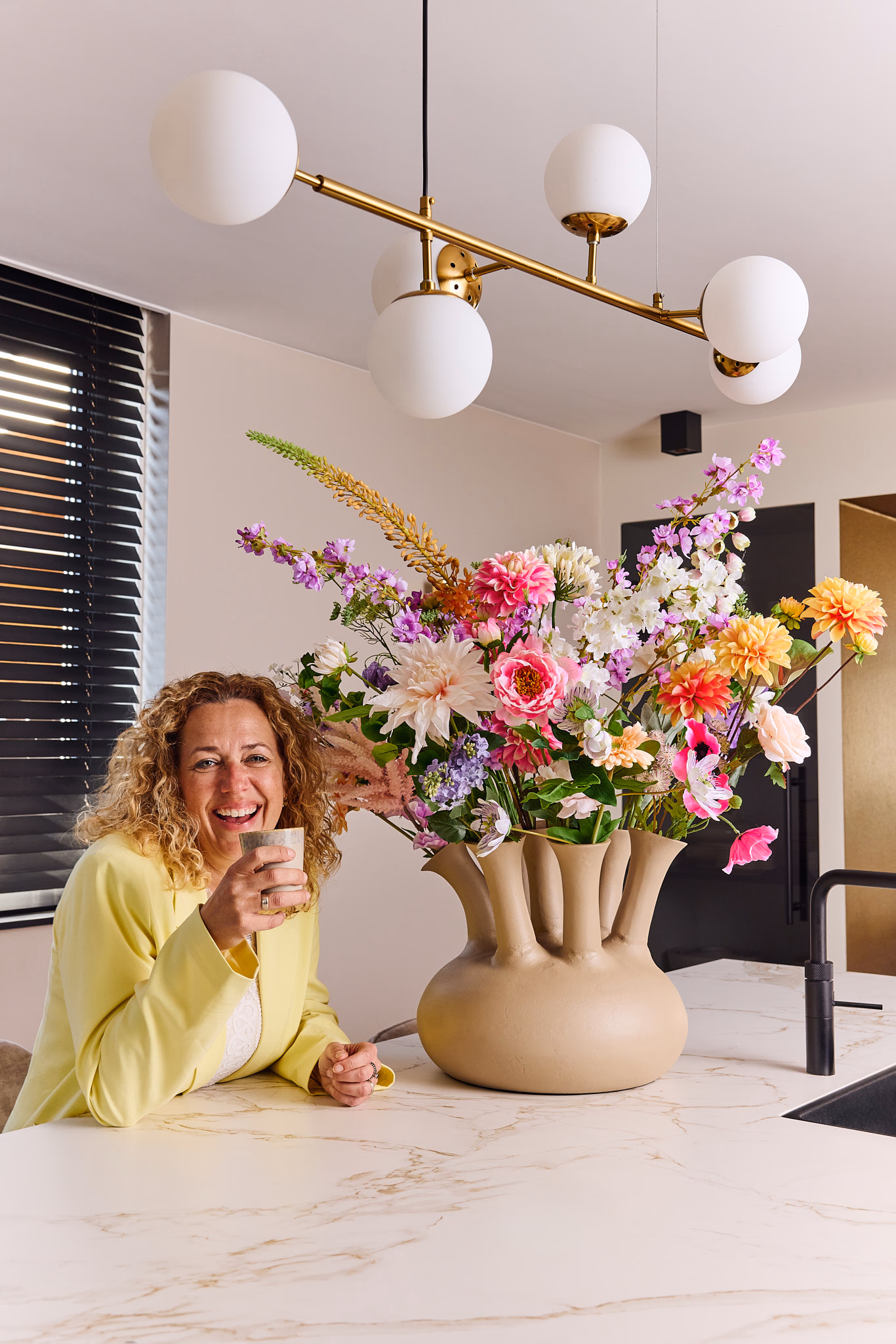 Vrouw in geel jasje glimlacht naast een groot luxe zijden bloemenboeket in een moderne beige vaas.
