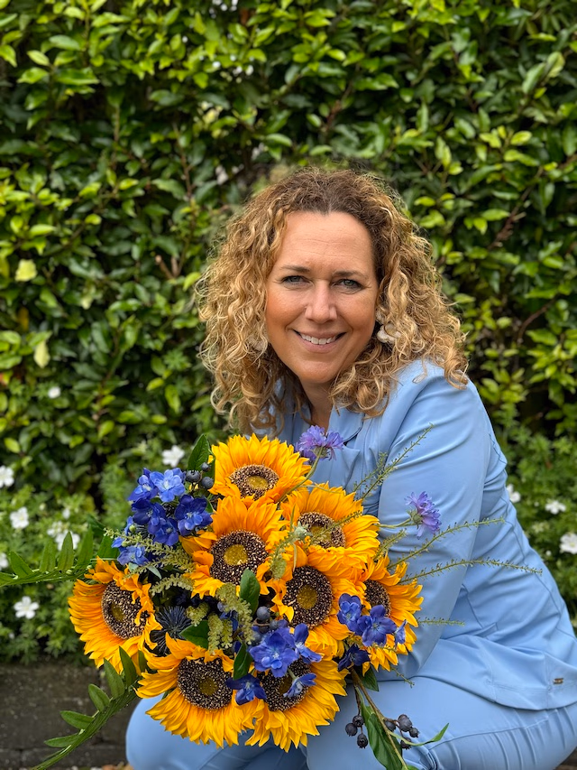 Een vrouw met krullend haar in een lichtblauw pak houdt een boeket van zonnebloemen en blauwe bloemen vast.